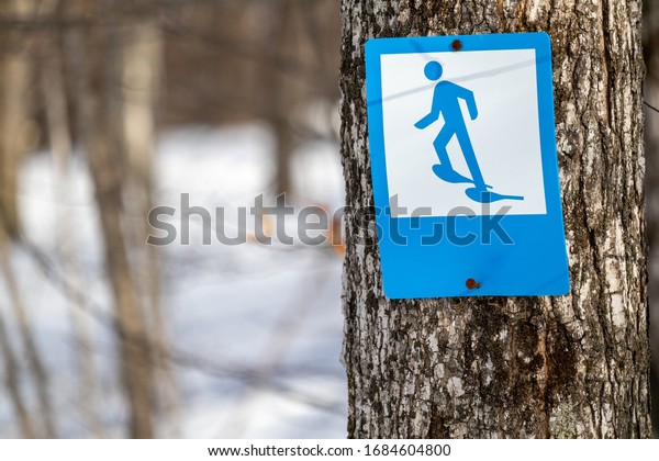 A blue-and-white sign\
features an illustration of a stick figure wearing snowshoes. This\
trail marker indicates a trail suitable for snowshoeing in the\
winter season.