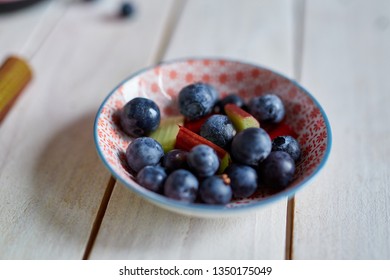 Blueberries with rhubarb in a bowl on a wooden table with rhubarb next to the bowl
