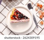 Blueberry cake slice served on a white plate with a bread cooling base, plus decoration in the from of blueberries, flower and additional props that add esthetics to the photo.