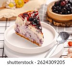 Blueberry cake slice served on a white plate with a bread cooling base, plus decoration in the from of blueberries, flower and additional props that add esthetics to the photo.