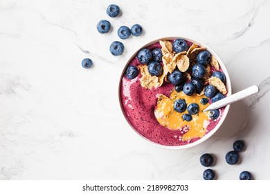 Blueberry smoothie bowl with peanut butter, cereal and fresh berries, white marble background, top view. Healthy vegan food concept.