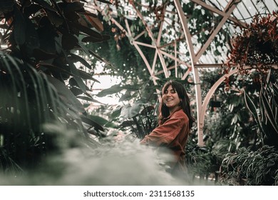 blue-eyed caucasian young woman wearing pretty earrings and red jacket smiling at camera inside beautiful botanic garden full of plants, botanic garden christchurch, new zealand - Shutterstock ID 2311568135