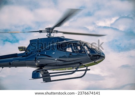 A blue-grey jet-powered helicopter hovering. High stratus clouds fill the sky with breaks showing blue skies. Rotors are motion blurred.