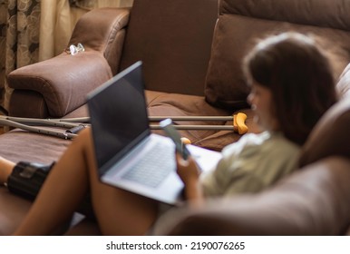 Bluered Asian Teen Girs With Injured Leg And Crutches Talking On The Phone And Laptop At Home, Sitting On The Sofa