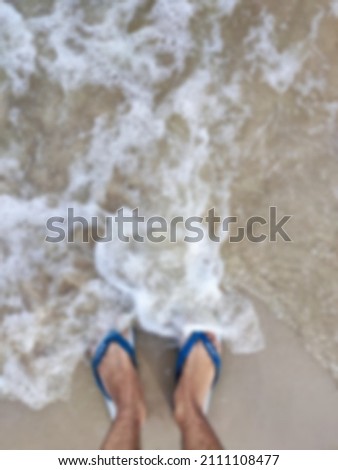 Blur background, man legs with blue sandals at sea foam waves on sand beach summer day
