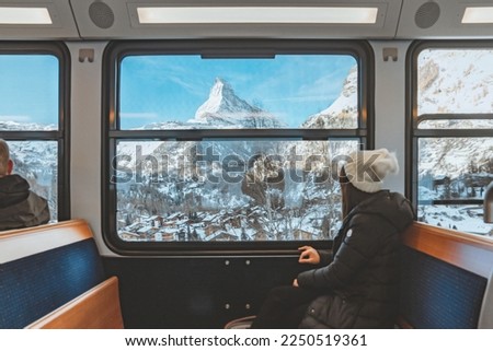 Blur foreground - young woman traveling looking out the window enjoying in Swiss Alps with the Matterhorn in winter background while sitting in the train. Tourist travel concept.