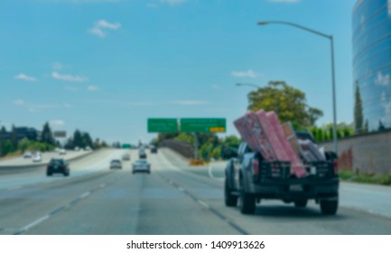 Blur. Pickup Truck With Open Tailgate Loaded With Mattresses And Home Furniture Drives In Slow Lane With Light Traffic On Highway To The New Home