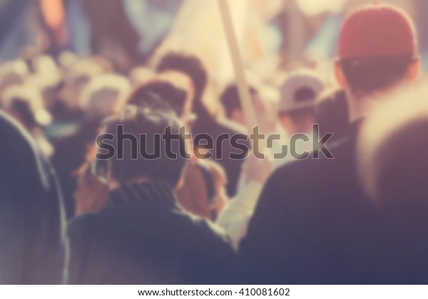 Blur unrecognizable\
crowd at political meeting, cheering audience looking at the stage\
and supporting political party, defocussed retro toned image with\
lens flare.