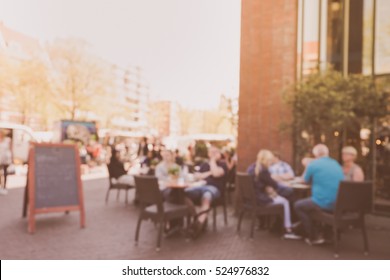 Blurred Abstract Background Of Outdoor Cafe Or Restaurant In Amsterdam. Outdoor Cafe With Tables, Chairs, Sign And Many People. For Create Montage Product Display In Golden Time.