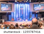Blurred abstract Christian people inside the church listening to preacher speaking. Defocused back view audience on row of raised sitting chairs looking at stage with large video projector screens