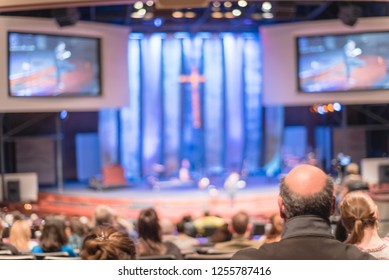 Blurred Abstract Christian People Inside The Church Listening To Preacher Speaking. Defocused Back View Audience On Row Of Raised Sitting Chairs Looking At Stage With Large Video Projector Screens