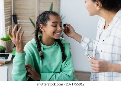 blurred african american woman doing makeup to cheerful teenage daughter