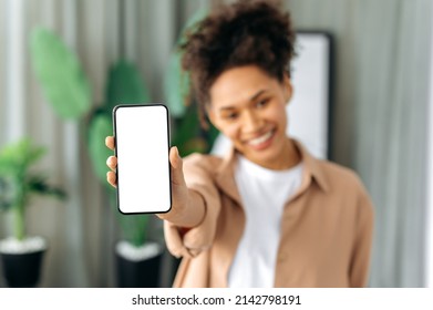 Blurred African American Young Woman In Casual Wear, Showing Modern Cell Phone With Blank White Screen, Stands At Home In A Living Room, Smiling, Mockup, Copy Space Concept