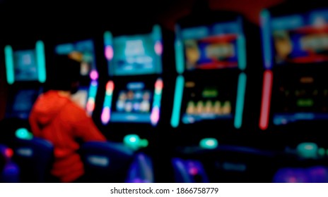 A blurred anonymized shot of a slot machine - video poker room, with lights glowing in the dark. A man, dressed in red, is playing at one machine. Soft pleasant bokeh.
 - Shutterstock ID 1866758779
