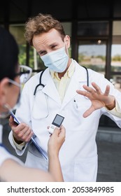 Blurred Asian Reporter Holding Voice Recorder Near Doctor In Medical Mask And White Coat Gesturing During Interview