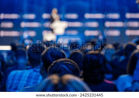 Blurred audience in focus watching a speaker at a conference with a blue background stage.