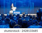 Blurred audience listening to a speaker at a professional conference event in a large auditorium.