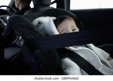 Blurred Baby In White Puffy Jacket Sit  In Rear-facing  Car Seat Beside Black Strap With Father Driving In The Background 