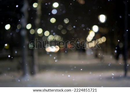 Blurred background. City view, lights, falling snow, night, street, bokeh spots of headlights of moving cars. Diffuse Urban backdrop winter scenery of street in city at night. Lantern light, snowfall