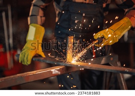 Blurred background of a worker in a work overall wearing a safety equipment and protective gloves while welding a metal construction. Glowing sparks flying all around the working facility.Copy space.