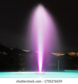 Blurred Bright Neon Purple Sprinkling Water Fountain Light Streaks, Pale Teal Neon Blue Green Swimming Pool Water In Empty Aqua Park Or Hotel Resort With No People At Warm Summer Night. Long Exposure