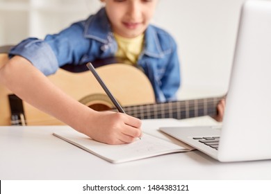 Blurred Child With Guitar Writing In Notebook During Online Music Lesson At Desk