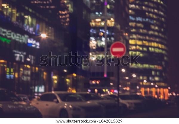 Blurred city\
background with No pass red sign. Defocused backdrop of glowing\
business buildings and parked\
cars