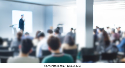 Blurred De-focused Audience In Conference Lecture Presentation Room. Corporate Presentations In Conference Hall. Seminar Speaker Giving Training To New Employees. Blurred  Hip Presenter Wearing Hat.