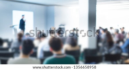Blurred De-focused Audience in Conference Lecture Presentation Room. Corporate Presentations in Conference Hall. Seminar Speaker Giving Training to New Employees. Blurred  Hip Presenter wearing Hat.
