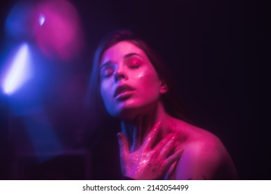 Blurred Fashionable Portrait Of A Woman With Glitter On Her Face And Bright Makeup In Neon Light Dancing At A Party. Defocused Woman In Purple Light With Glare On A Dark Background.