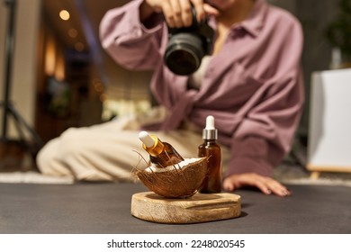Blurred female photographer taking photo of bottles with oil and coconut on wooden board on camera at studio. Beauty and body care content for photostocks, commerce, social networks and advertising - Shutterstock ID 2248020545
