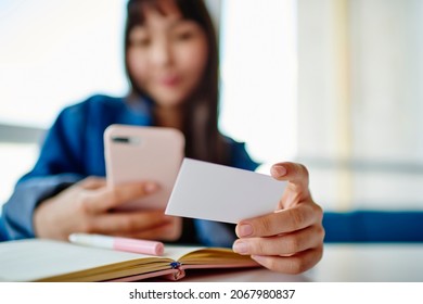 Blurred Female Student With Education Textbook Holding White Business Card And Using Smartphone Technology For Dialing Number, Woman Browsing Mobile Website While Checking Contact Information