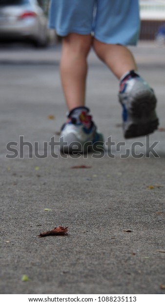 Blurred focus\
 the boy is run on a concrete road.Low angle view picture shows the\
legs of children wear run shoes and blue shorts.on the street there\
are flowers wither,car is\
blur