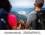 Blurred foreground close up from behind of female and male hikers with backpack and hike-sticks admiring panorama with mountain lake