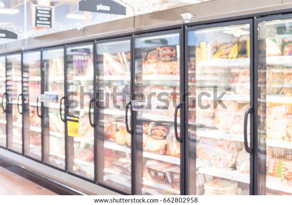Blurred frozen food section at retail store\
in America. Huge glass door aisle with variety pack of processed\
pizza. Freezer full assortment of frozen pizza in local\
supermarket, defocused\
background.