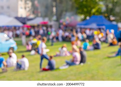 Blurred Group Of People, Family Members Enjoy Outdoor Picnic And Festival, Open-air Free Concert In Public Park. Audience Sits On Grass