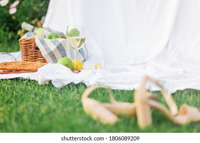 Blurred High-heeled Shoes Lay Beside Wicker Basket With Set For Picnic. On Green Grass And White Sheets