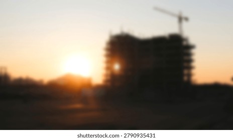 A blurred image of a building under construction with a crane silhouetted against a vibrant sunset. The sun is setting behind the building.