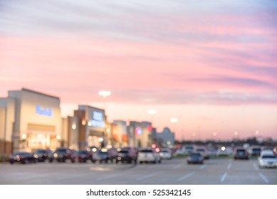 Blurred Image Exterior View Of Modern Shopping Center In Humble, Texas, US At Sunset. Mall Complex With Row Of Cars In Outdoor Uncovered Parking Lots And Bokeh Light Of Retail Store In Background.