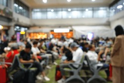 A Blurred Image Of A Group Of People Waiting In The Airport For Their Flight There Are Children, Adults, Women, And Men. Doing Their Own Thing. Some Are Reading And Playing On Their Mobile Phones.