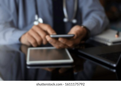 Blurred Image Of Male Doctor In Blue Shirt With Tie, Hand Holding And Using Mobile Smart Phone With Modern Laptop Computer, Medical Stethoscope And Wireless Mouse On The Desk. Medical Online Concept.