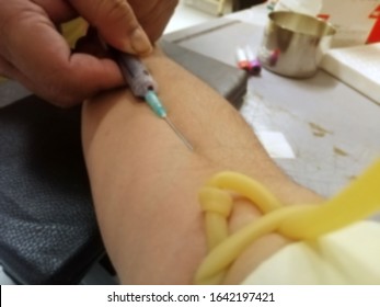 Blurred Image Of Nurse Pricking Needle Syringe In The Arm Patient Drawing Blood Sample For Blood Test, Puncture Of A Vein Through The Skin In Order To Withdraw Blood For Analysis.