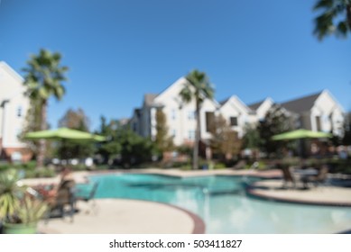 Blurred Image Outdoor Swimming Pool And Woven Rattan Pool Beds With Modern Apartment Houses In Background. Apartment Backyard Landscape With Swimming Pool, Palm Trees And Clear Blue Sky In Texas, US.