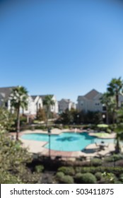 Blurred Image Outdoor Swimming Pool And Woven Rattan Pool Beds With Modern Houses In Background. Elevated View Apartment Backyard Landscape With Swimming Pool, Palm Trees, Clear Blue Sky In Texas, US.