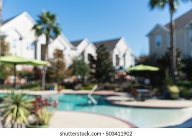 Blurred Image Outdoor Swimming Pool And Woven Rattan Pool Beds With Modern Apartment Houses In Background. Apartment Backyard Landscape With Swimming Pool, Palm Trees And Clear Blue Sky In Texas, US.