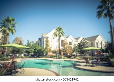 Blurred Image Outdoor Swimming Pool And Woven Rattan Pool Beds With Modern Apartment Houses In Background. Apartment Backyard Landscape With Pool, Palm Trees, Clear Blue Sky, Texas, US. Vintage Filter