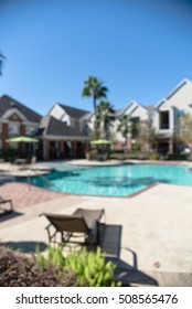 Blurred Image Outdoor Swimming Pool And Woven Rattan Pool Beds With Modern Apartment Houses In Background. Apartment Backyard Landscape With Swimming Pool, Palm Trees And Clear Blue Sky In Texas, US.