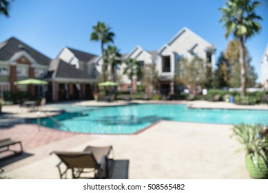 Blurred Image Outdoor Swimming Pool And Woven Rattan Pool Beds With Modern Apartment Houses In Background. Apartment Backyard Landscape With Swimming Pool, Palm Trees And Clear Blue Sky In Texas, US.