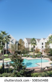 Blurred Image Outdoor Swimming Pool And Woven Rattan Pool Beds With Modern Houses In Background. Elevated View Apartment Backyard Landscape With Swimming Pool, Palm Trees, Clear Blue Sky In Texas, US.