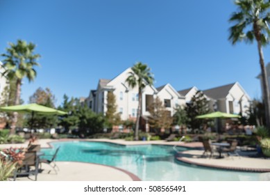 Blurred Image Outdoor Swimming Pool And Woven Rattan Pool Beds With Modern Apartment Houses In Background. Apartment Backyard Landscape With Swimming Pool, Palm Trees And Clear Blue Sky In Texas, US.